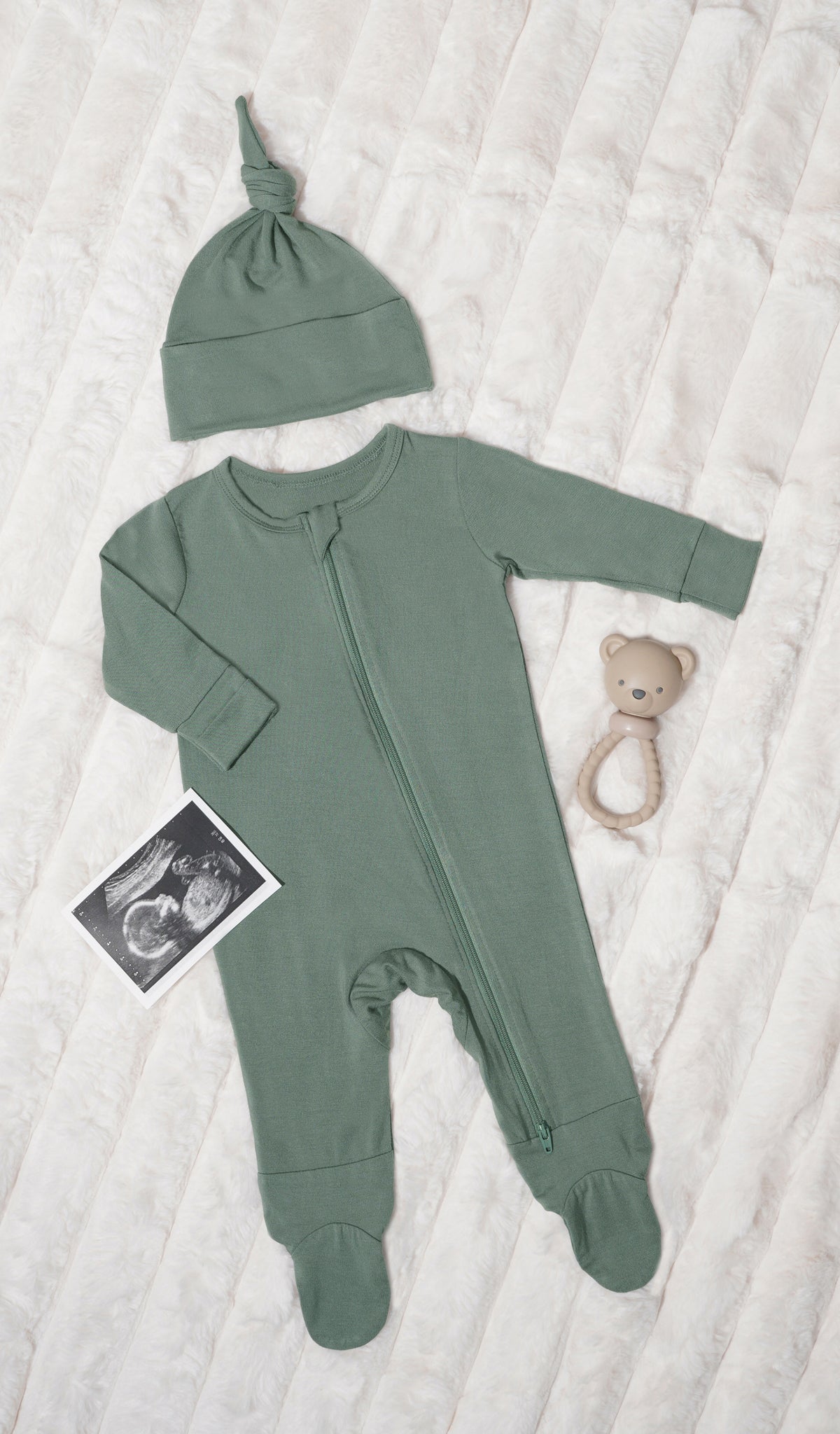 Basil Footie 2-Piece. Long sleeve footie with zip front, matching knotted baby hat, ultrasound picture and a rattle on soft beige blanket.