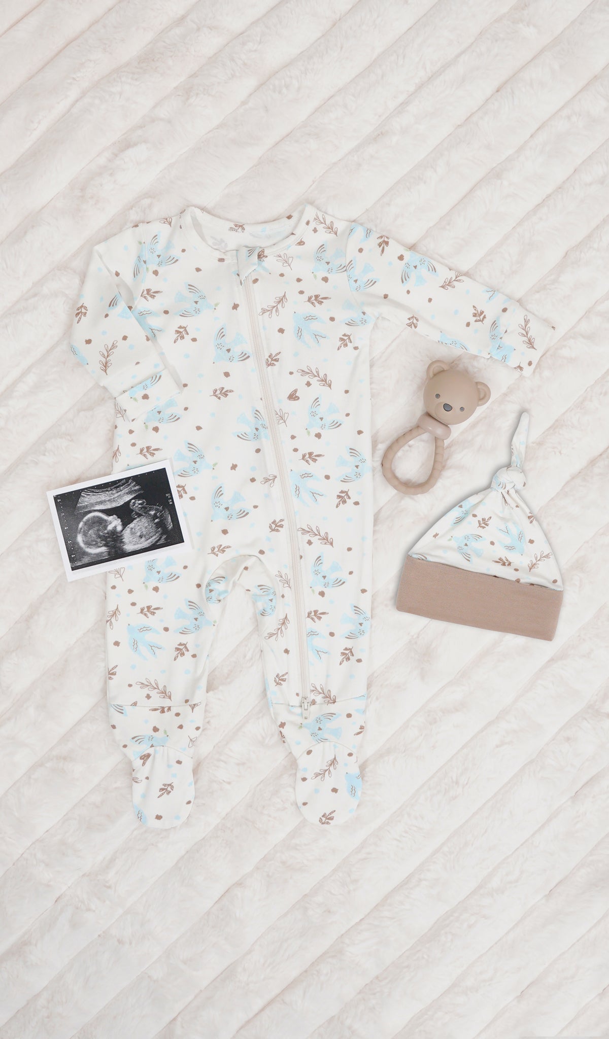 Nest Footie 2-Piece. Long sleeve footie with zip front, matching knotted baby hat, ultrasound picture and a rattle on soft beige blanket.