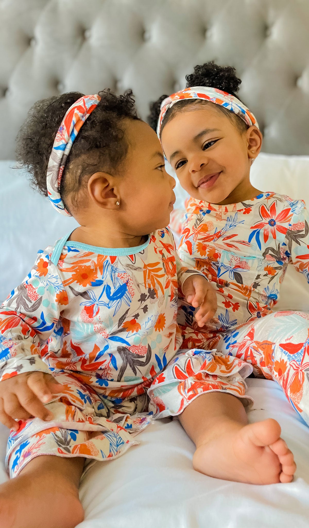 Posy Ruffle Romper 2-Piece on baby girl wearing matching headwrap while sitting and looking back at big sister wearing long sleeve top, pant and matching headwrap from Charlie 3-Piece PJ set.