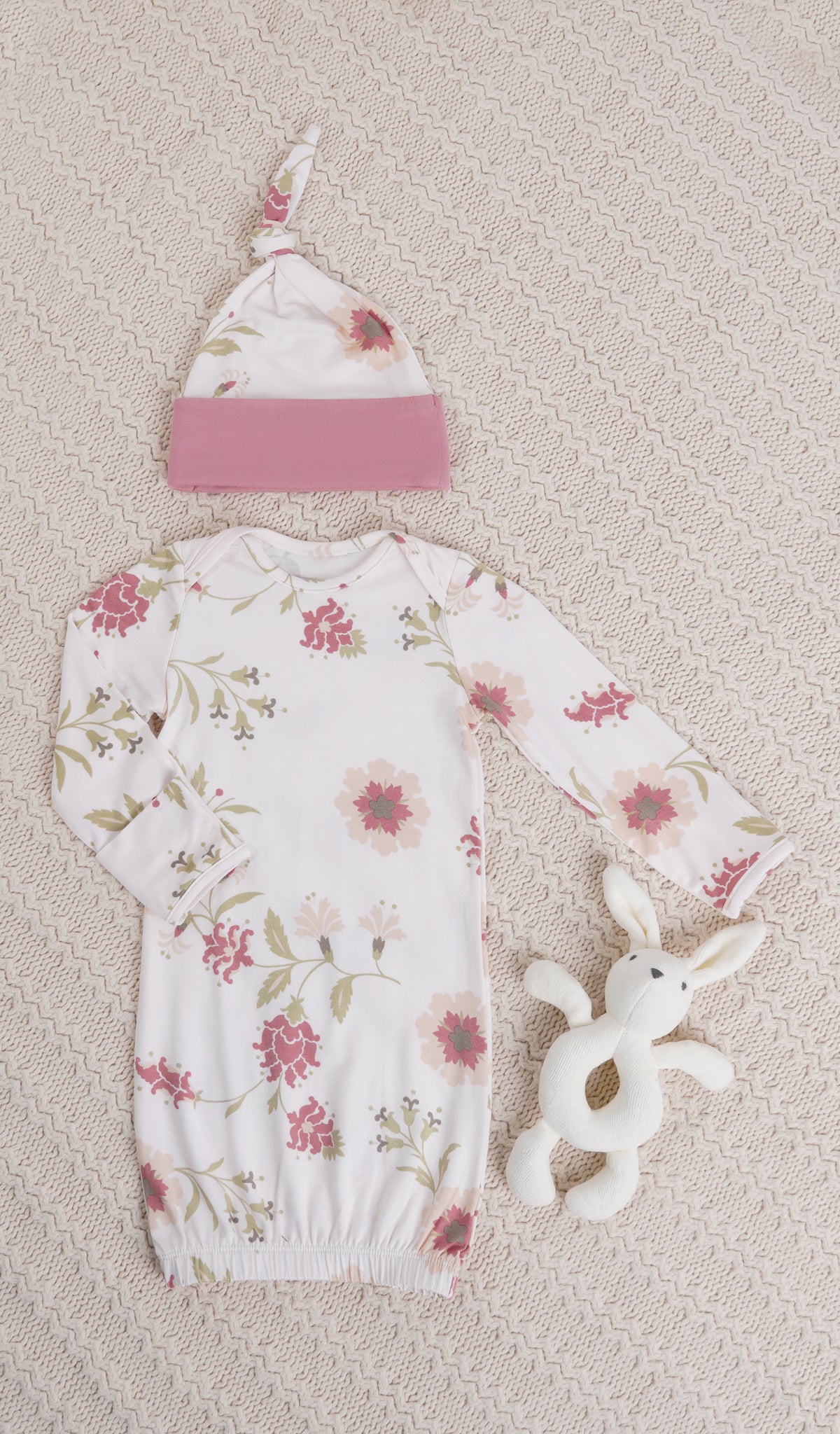 Cottage Floral Analise 5-Piece Set, gown and knotted hat for baby with bunny rattle on a textured beige blanket.