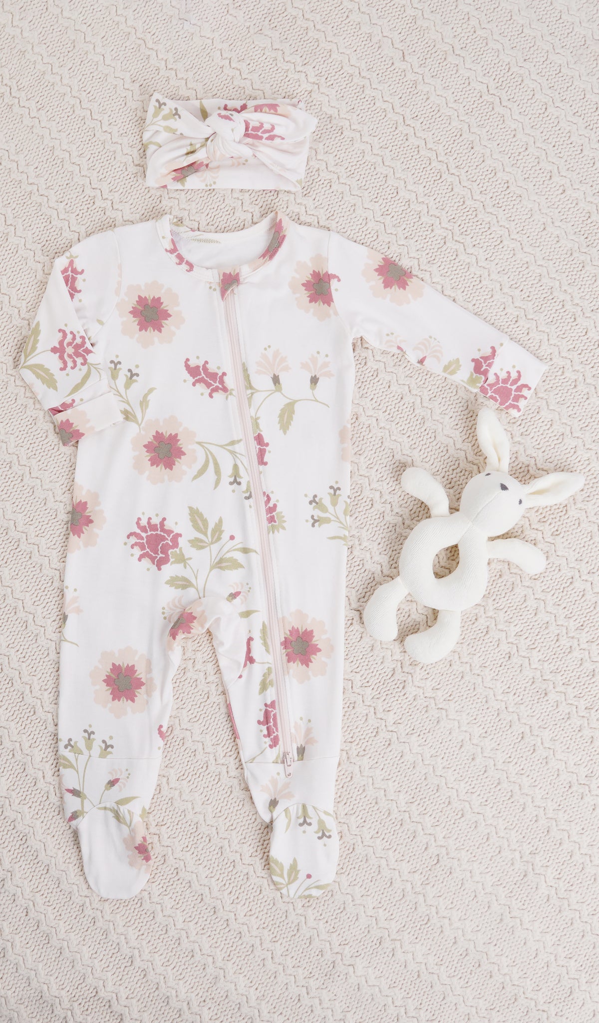 Cottage Floral Footie 2-Piece Set. Zip front footie for baby with matching headwrap and a bunny rattle on knitted beige blanket.