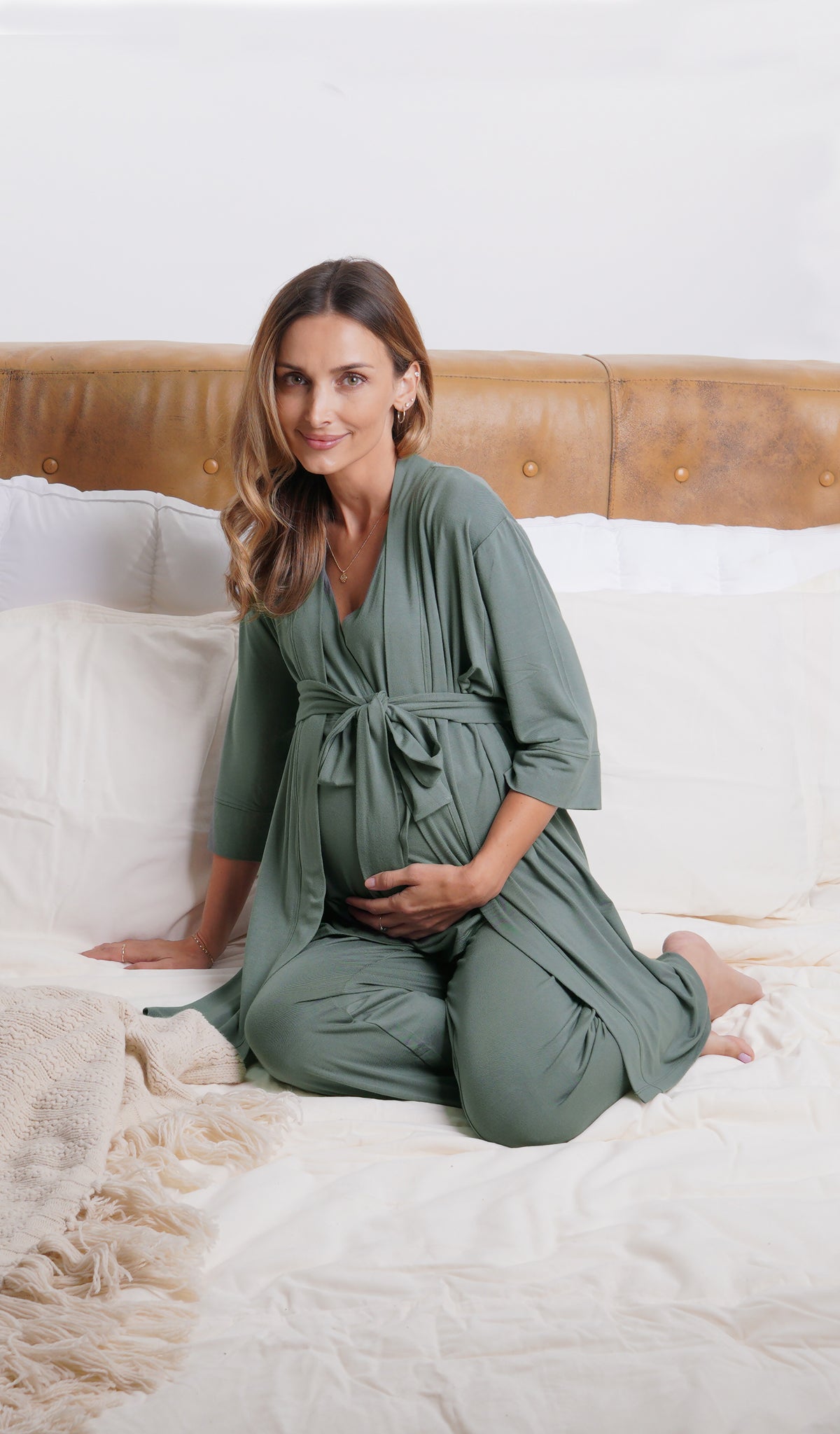 Fog Analise 3-Piece Set, woman wearing robe, tank top and pant sitting on bed leaning back on one hand while holding belly with other hand.