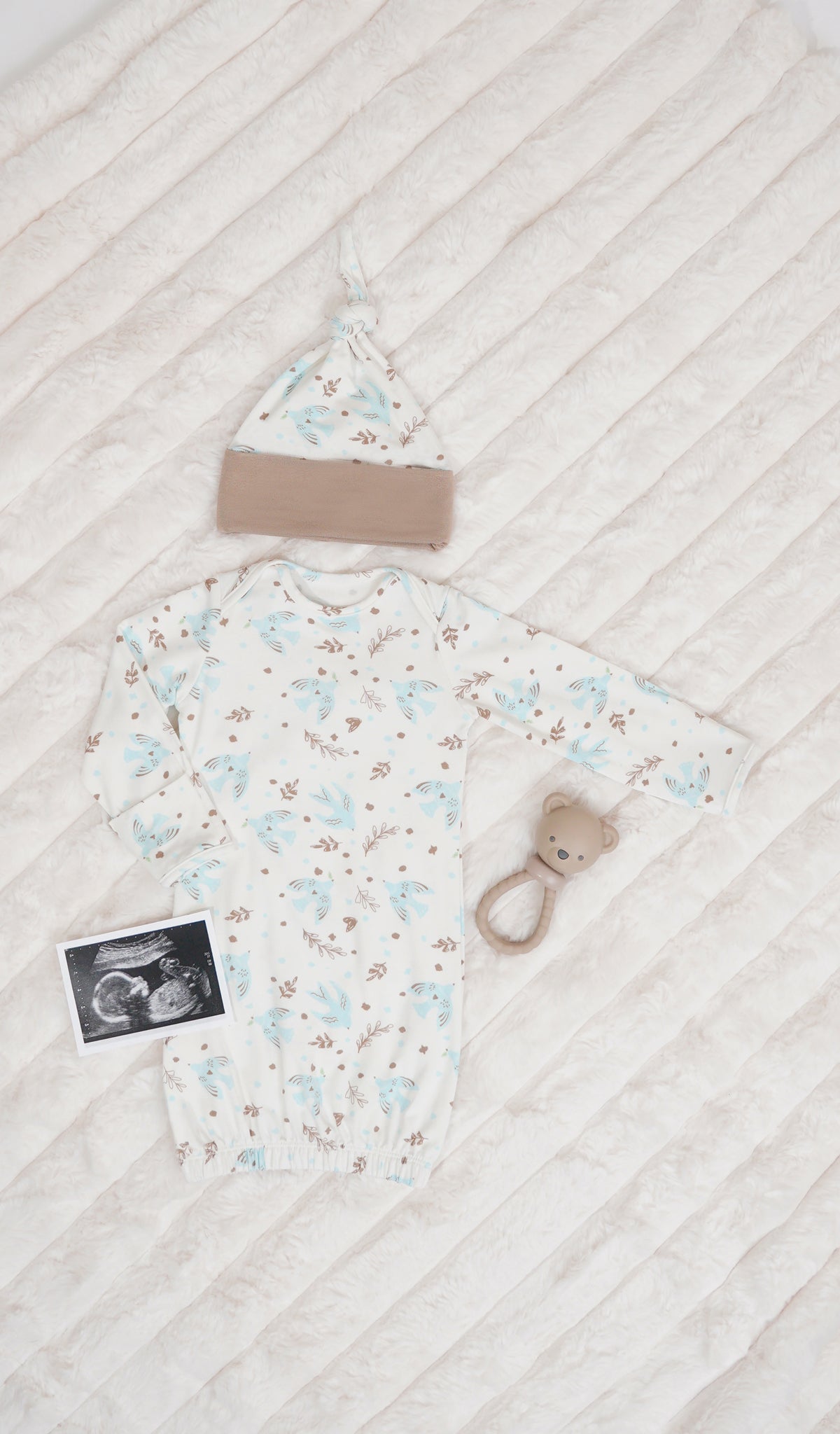 Nest Analise 5-Piece Set, gown and knotted hat for baby with rattle and ultrasound image on a textured beige blanket.